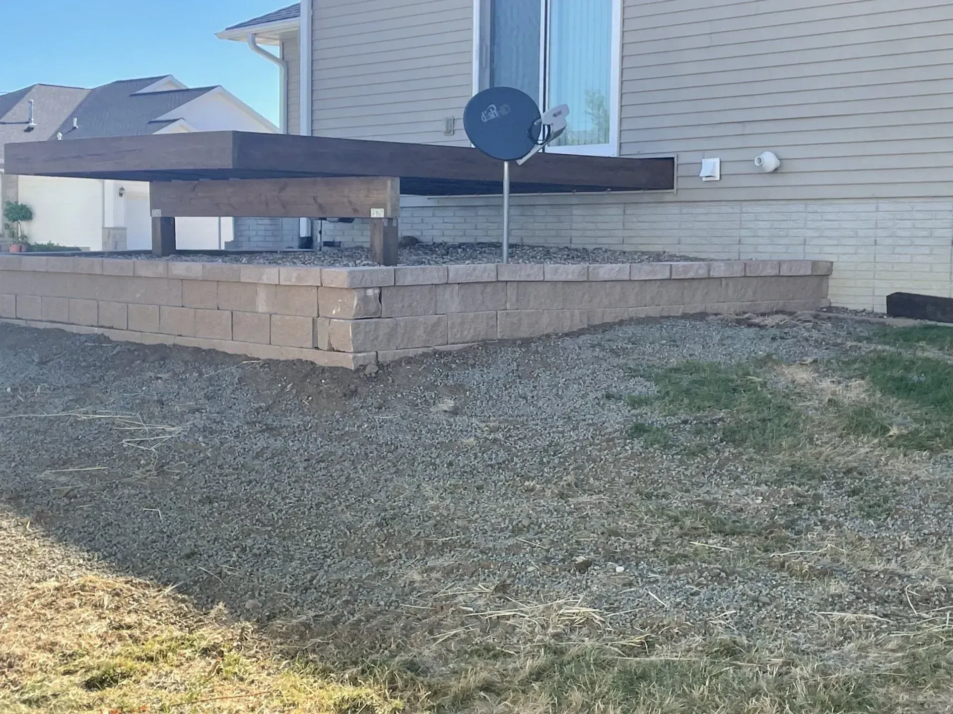 Raised wooden deck attached to a beige house, with a retaining wall and gravel yard.