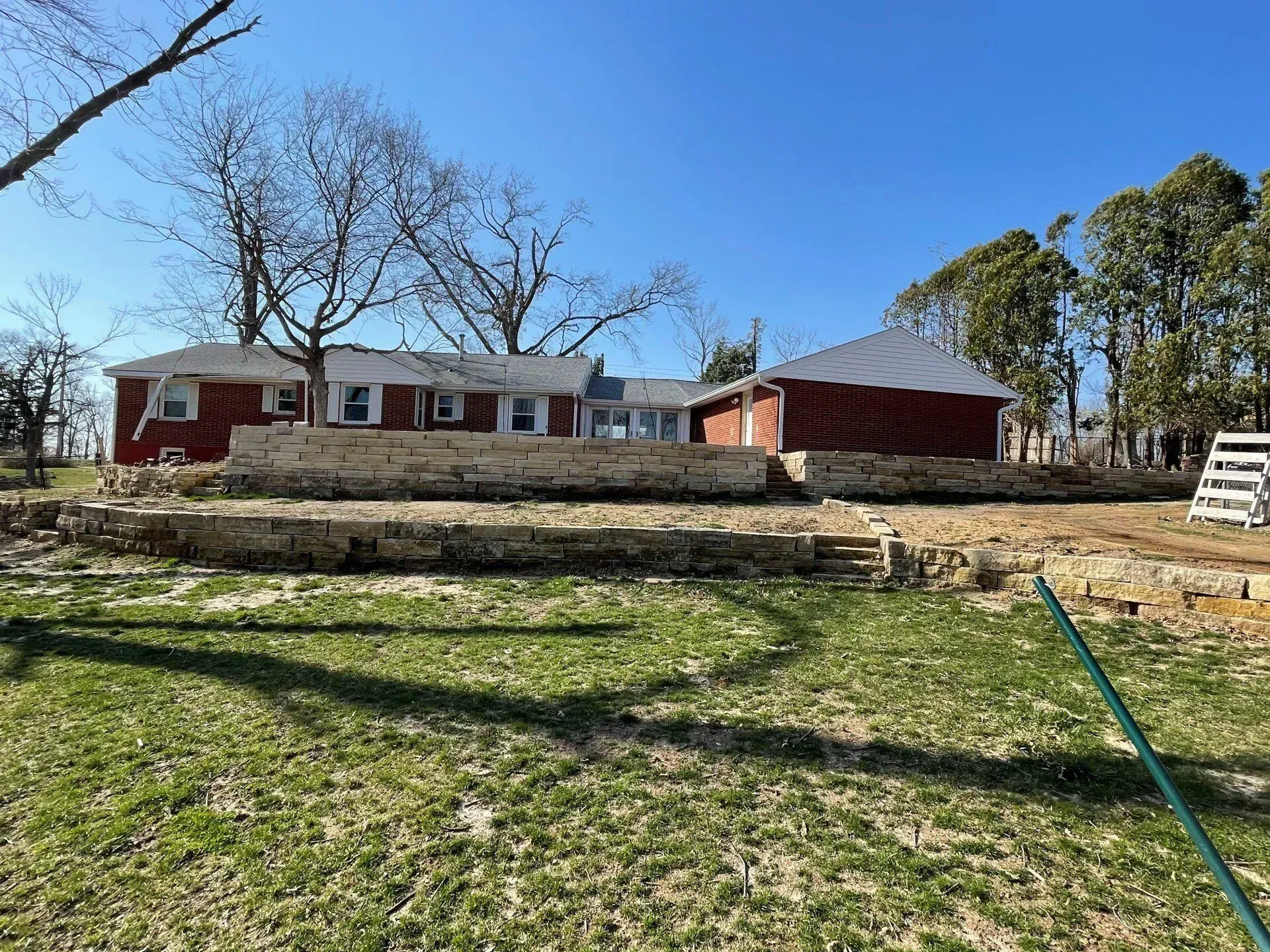 Red brick house and shed with stone retaining walls on a grassy hill under a blue sky.