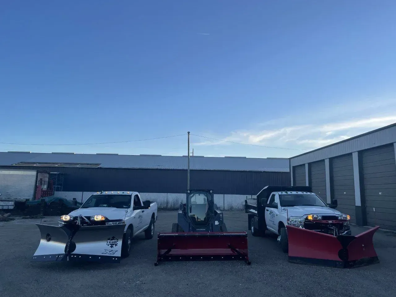 Three white snowplows and a small tractor, parked outside with a blue sky.
