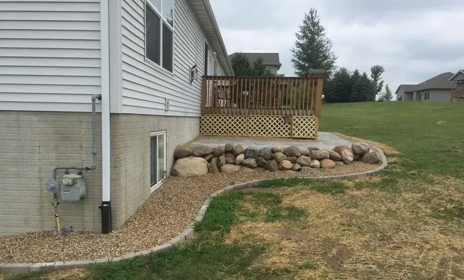 Back of a house with a wooden deck, a stone retaining wall, and a gravel path in a grassy yard.